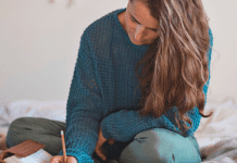 Woman dressed in pants and a sweater sitting on top of her bed writing in her journal