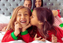 Two sisters in holiday pajamas laying on a bed with their parents in the background